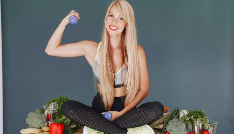 Woman lifting a small dumbbell while surrounded by fresh vegetables, embodying health and fitness.